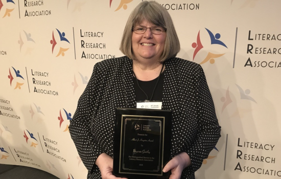 A woman with chin-length gray hair and glasses wears a polka dot blazer and holds a plaque. The wall behind her has repeating logos next to the words “Literacy Research Association.”