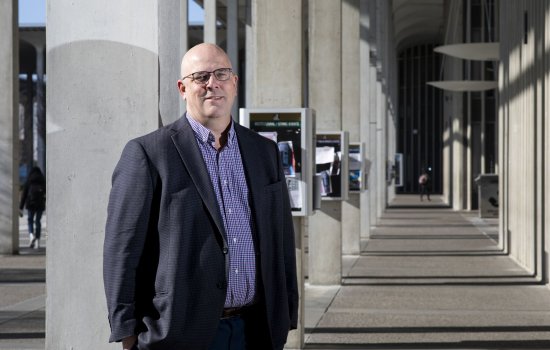 Shawn Bushway stands on the pillared UAlbany Podium in a blue jacket and purple button-doown shirt