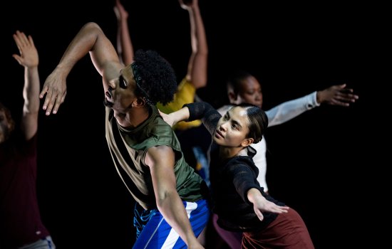 two dancers in foreground lean sideways in connection to one another; arms of other dancers are visible in background 
