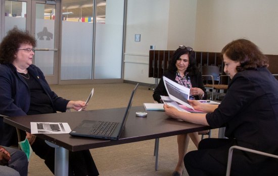 Three women sit around a table with a laptop on it, looking at printed materials