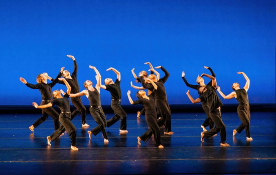 many dancers costumed in black lean back looking upward with arms extended high