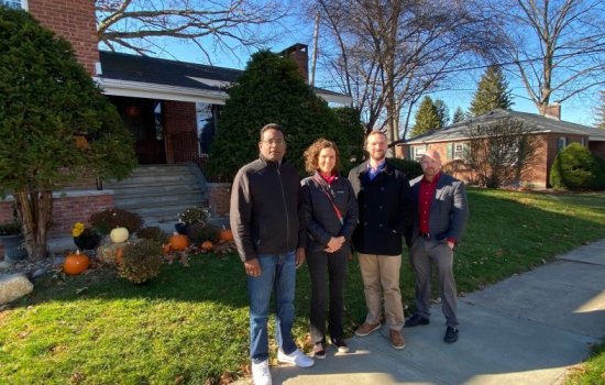Four people in jackets stand on a sidewalk outside a brick house with a lawn decorated pumpkins.