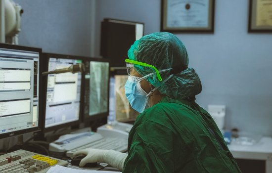 A healthcare worker wearing scrubs, goggles, mask and gloves, works at bank of computers in a dimly-lit room. 
