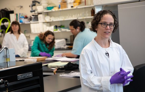 A woman with short brown curly hair and glasses wears a white lab coat and purple latex gloves, her hands clasped together as she smiles and speaks to someone outside the frame.