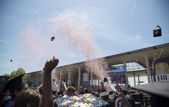 A confetti cannon shoots a blast of purple and gold confetti over a sea of black mortar board hats and hands raised in celebration at UAlbany's spring 2023 Undergraduate Commencement.