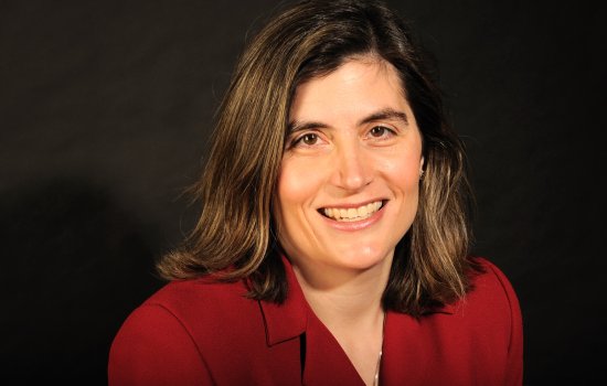 Portrait of Erin Bell; Erin is smiling wearing a red blazer with a dark brown backdrop