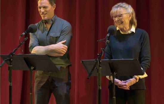 two smiling actors stand side by side and in front of music stands and microphones