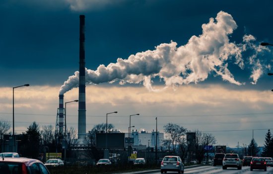 Landscape image of factory smokestacks in the background and a busy multi-lane highway in the foreground. The top layer of the sky is blue; the layer closer to the ground is cloudy with a light haze. 