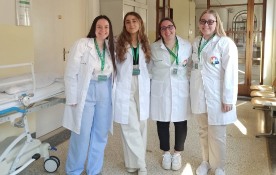Four female students wearing white medical jackets and green lanyards with nametags stand in a sunlit hospital hallway. All are smiling. Part of a stretcher is visible at left. 