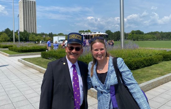 President Havidán Rodríguez and Jessica Castner, both smiling broadly, pose together in front of UAlbany's new electric trolley. It is a beautiful, sunny summer day. The background landscape is full of green grass, full green trees, and planters with boxwood bushes and purple flowers.