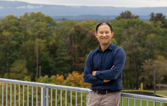 A man with short black hair in a navy shirt and tan pants leans against an outdoor railing, arms crossed, and smiles for a picture. A vast landscape of trees and hills can be seen behind him.