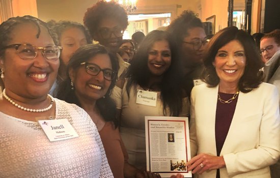 Governor Kathy Hochul holds the WGSS newsletter while surrounded by smiling WGSS faculty and students in a hallway of the Governor's Mansion.