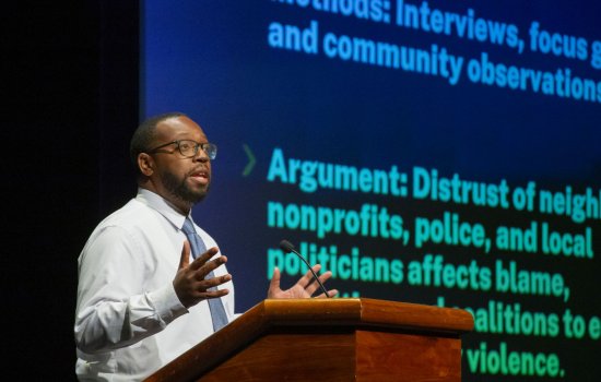A man in glasses wearing a shirt and tie stands at a podium in front of a projection of words including "distrust, nonprofits, police and local politicians"