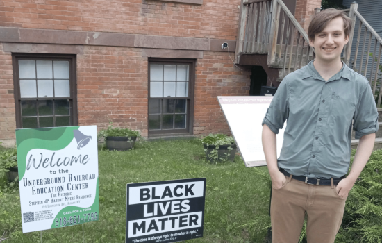 A young man with light brown hair in a button down shirt and tan pants stands in front of a brick building next to a "Black Lives Matter" sign and sign that reads "Welcome to the Underground Railroad Education Center."