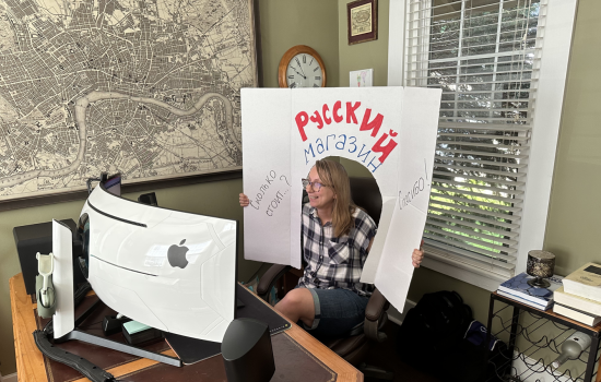 A woman, Maria Goebert, is seated in front of a computer screen in an office. She smiles and holds a white cardboard cutout featuring Russian phrases drawn on in marker over her head.