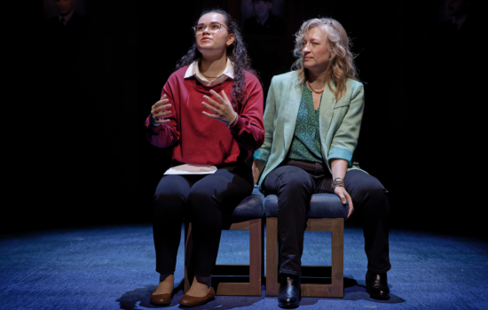 Two women are seated next to each other on a stage. The woman on the left, in a red sweater over a white button down shirt, gestures with her hands while staring in the distance as the other woman, in a green blouse and blazer, watches her.