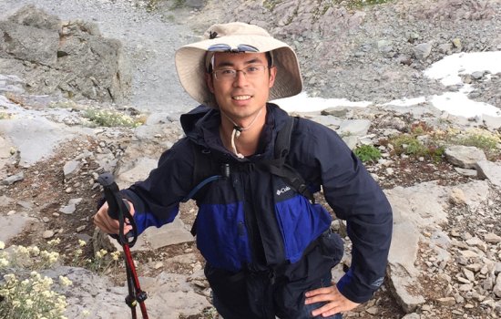 Felix Ye poses on a rocky trail while wearing glasses, a blue Columbia jacket, a hat and holding two hiking poles..