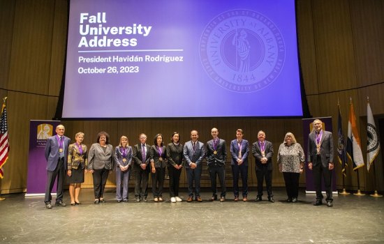 A line of 13 people standing shoulder to shoulder on a stage in front of a blue screen that reads: "Fall University Address, President Havidan Rodriguez, October 26, 2023."