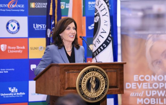 Gov. Hochul speaks from a wooden podium with the University at Albany seal, in front of a colorful poster of SUNY college logos
