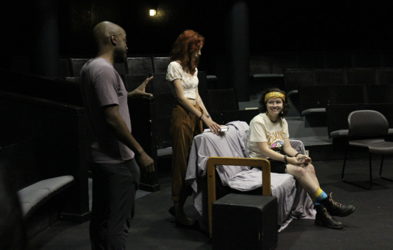 Three people appear in frame. A man stands to one side gesturing to a woman who is seated, as another woman stands directly behind her watching.