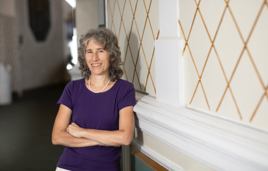 A woman with shoulder-length gray hair wears a purple t-shirt and smiles for a portrait with her arms crossed.