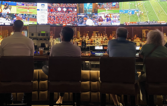 Four people are seated at a dimly-lit bar watching sports on a row of TV screens