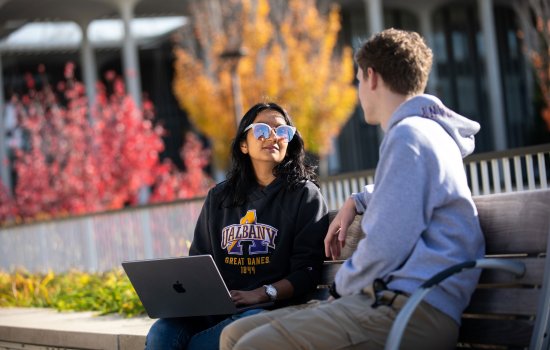 Two students wearing sweatshirts sit on a bench on a sunny fall day. One holds a laptop on their lap.