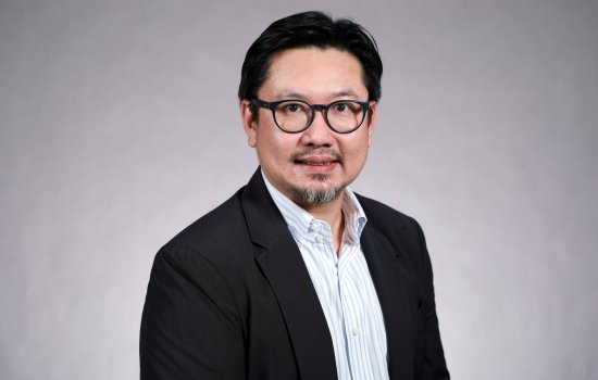 Portrait of Ricky Leung wearing glasses, a black blazer and a white, striped button-down shirt, seated in front of a gray background.