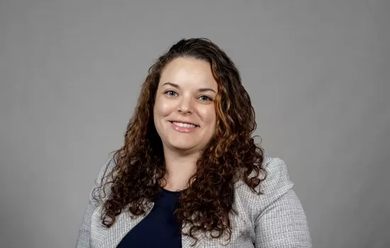 Sarah Domoff poses for a portrait against a gray background wearing a light gray jacket over a navy blue shirt. Domoff is smiling and has brown curly hair. 