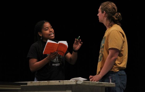 A woman holds a red book and gestures with a pen to a taller man facing her as the two rehearse for a play.
