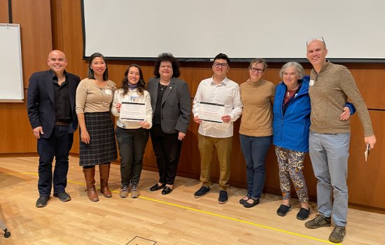 From left to right, Andrew Poulos, Cheryl Andam, Manuela Montoya Giraldo, Jeanette Altarriba, Andrew Munoz Gamba, Cara Pager, Marlene Belfort and Ken Halvorsen pose and smile in a conference room. Manuela and Andrew both hold certificates.