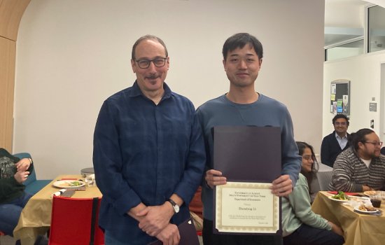 A man wearing a plain slate gray shirt holds a certificate and smiles as he stands on the right side of the image, next to man in a blue plaid button up and glasses.