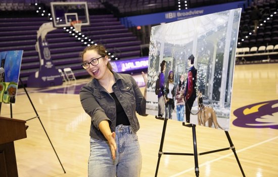 A woman stands on a basketball court smiling and gesturing toward an easel with a digital drawing of students standing in the snow on the UAlbany campus with a leashed Great Dane.