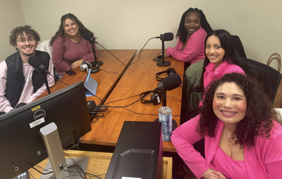 Five young people dressed in pink sit around a table topped with microphones, headphones and a Dell computer. They smile at the camera. 