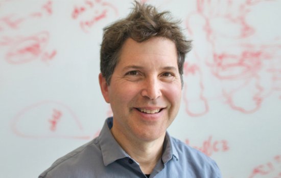 David Baker stands and smiles in front of an out-of-focus white board with red writing on it.