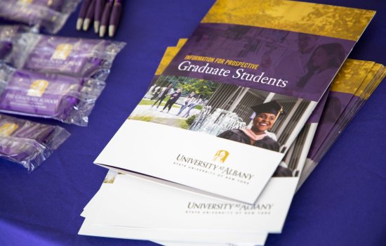 A pile of brochures reading "Information for prospective Graduate Students, University at Albany" on a purple tablecloth