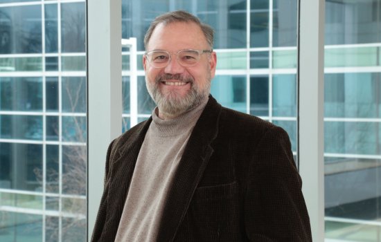 Portrait of Scott Tenenbaum, who is wearing clear-rimmed glasses, a dark brown blazer and tan turtleneck sweater. Tenenbaum is smiling, standing in front of a wall of glass windows.