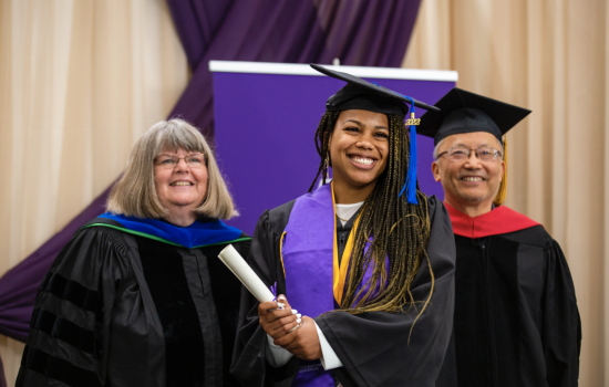 A smiling graduate in a black cap and gown holds a diploma as she poses for a portrait on stage with two others dressed in black graduation robes.