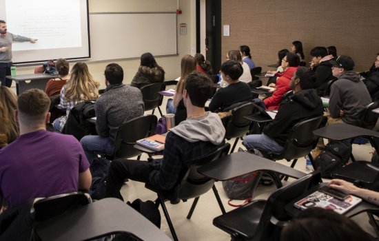 Man in gray sweater points to a projection as he speaks to a classroom full of students.