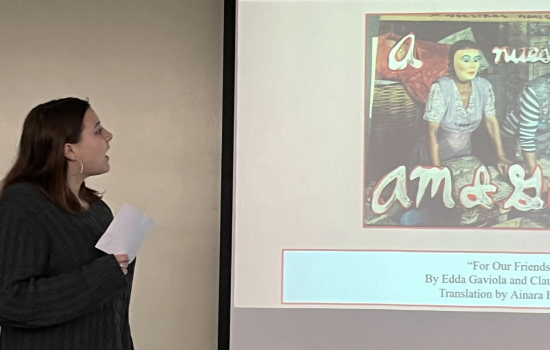 A young woman with shoulder length brown hair and hoop earrings speaks at the front of a room next to a screen depicting a woman in a dress and plastic face mask.