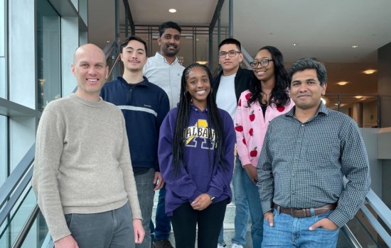 Six people pose together for a group portrait on an open stairwell in the RNA Institute atrium. There are five men and two women. All are smiling.