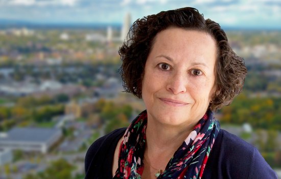 Portrait of Jean Moore wearing a navy top, red and blue flower printed scarf and gold necklace with a small green pendant. Jean has short brown hair and is smiling. The background is blurred and depicts a view of the Albany skyline.