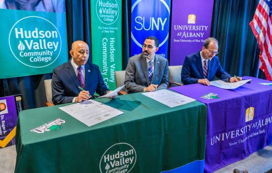 Three men in suits sign oversized pieces of paper at a table with a gree Hudson Valley Community College tablecloth on one side and a purple University at Albany tablecloth on the other. HVCC, SUNY and UAlbany banners are behind them.