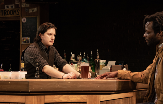 A man in a pinstripe shirt and vest stands behind a wooden bar and looks at a customer seated at the bar. A tall glass of beer sits between them.
