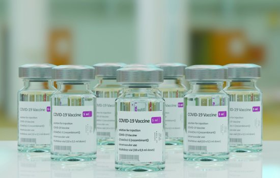 Image features seven COVID-19 vaccine vials with silver lids, arranged on a sleek, white tabletop. The background is blurred.