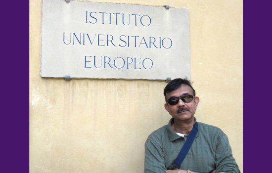 Kajal Lahiri stands with his arms crossed wearing a green long sleeved shirt and sunglasses. A blue bag strap falls across his chest and the bottom of the sign on the wall behind him reads "Instituto Universitario Europeo."