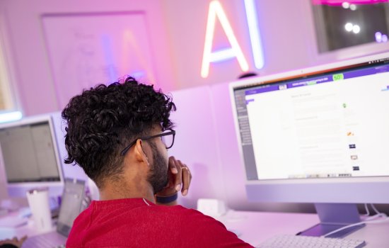 A student views a computer screen in front of a wall with a neon sign that says "AI." 