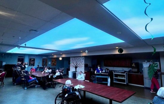 People in chairs and wheelchairs sit around tables under what appears to be a large window in the ceiling, showing blue sky and whispy clouds.
