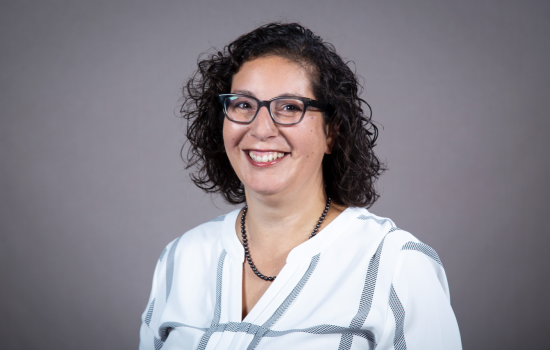 A woman with shoulder-length curly brown hair and glasses wears a striped blouse and smiles for a portrait against a gray backdrop.