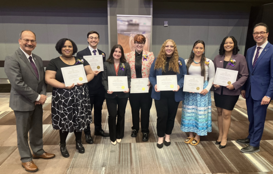 Seven students in professional attire stand holding award certificates and smile for a picture with UAlbany President Havidán Rodríguez and Vice President for Student Affairs Michael Christakis in a conference room.